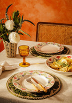 Dining table setting with floral plates, glasses, and a bowl of pasta against an orange wall.