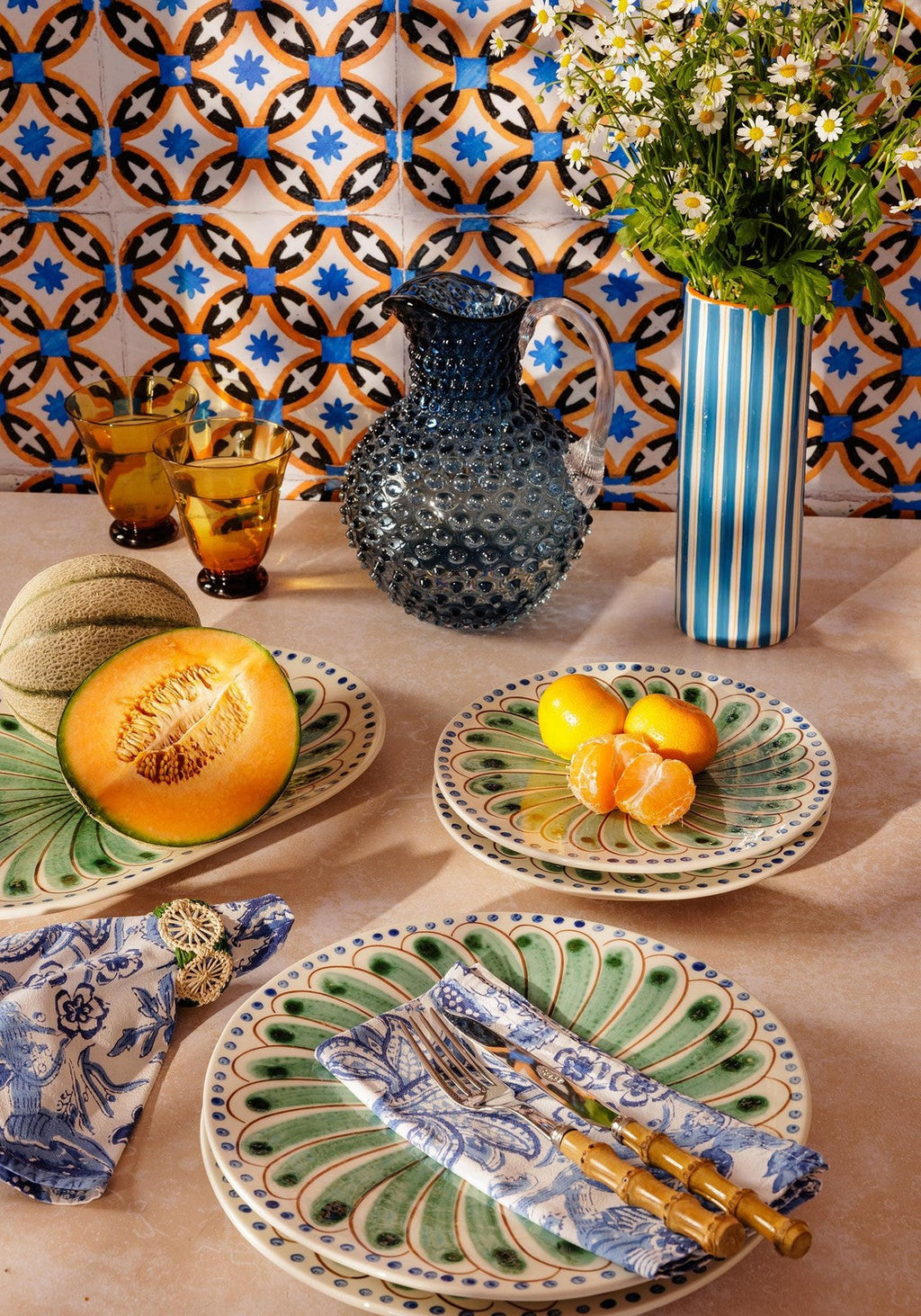 Decorative table setting with ceramic plates, a pitcher, and fruit against a patterned tile wall.