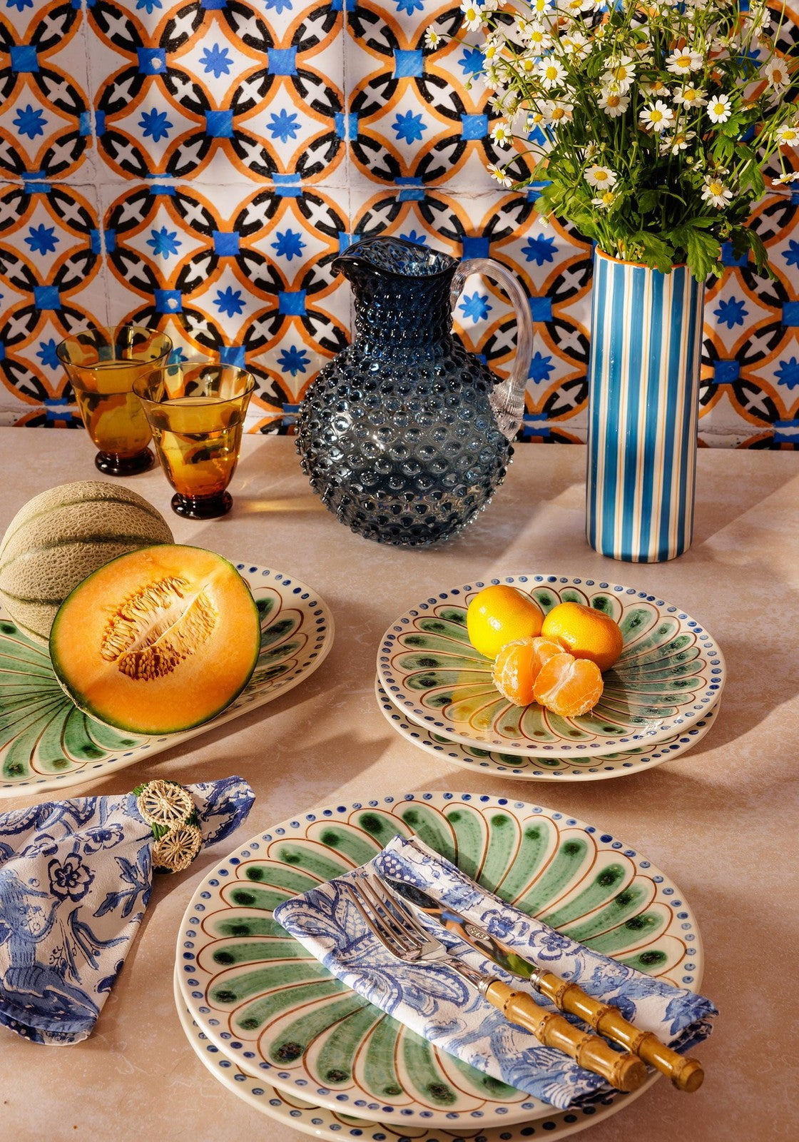 Decorative table setting with ceramic plates, a pitcher, and fruit against a patterned tile wall.