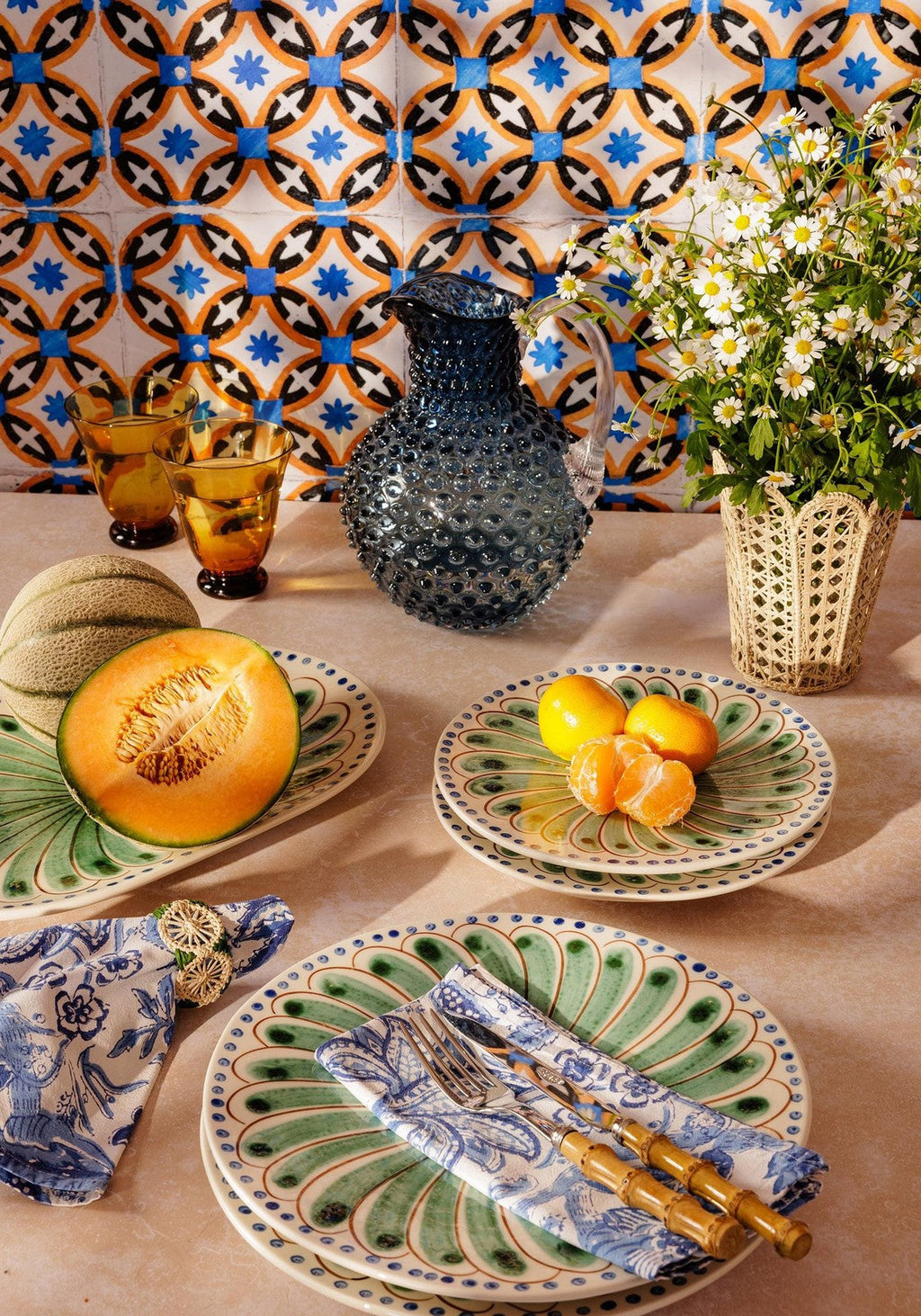 Decorative table setting with patterned plates, fruit, and a vase against a colorful tiled wall.