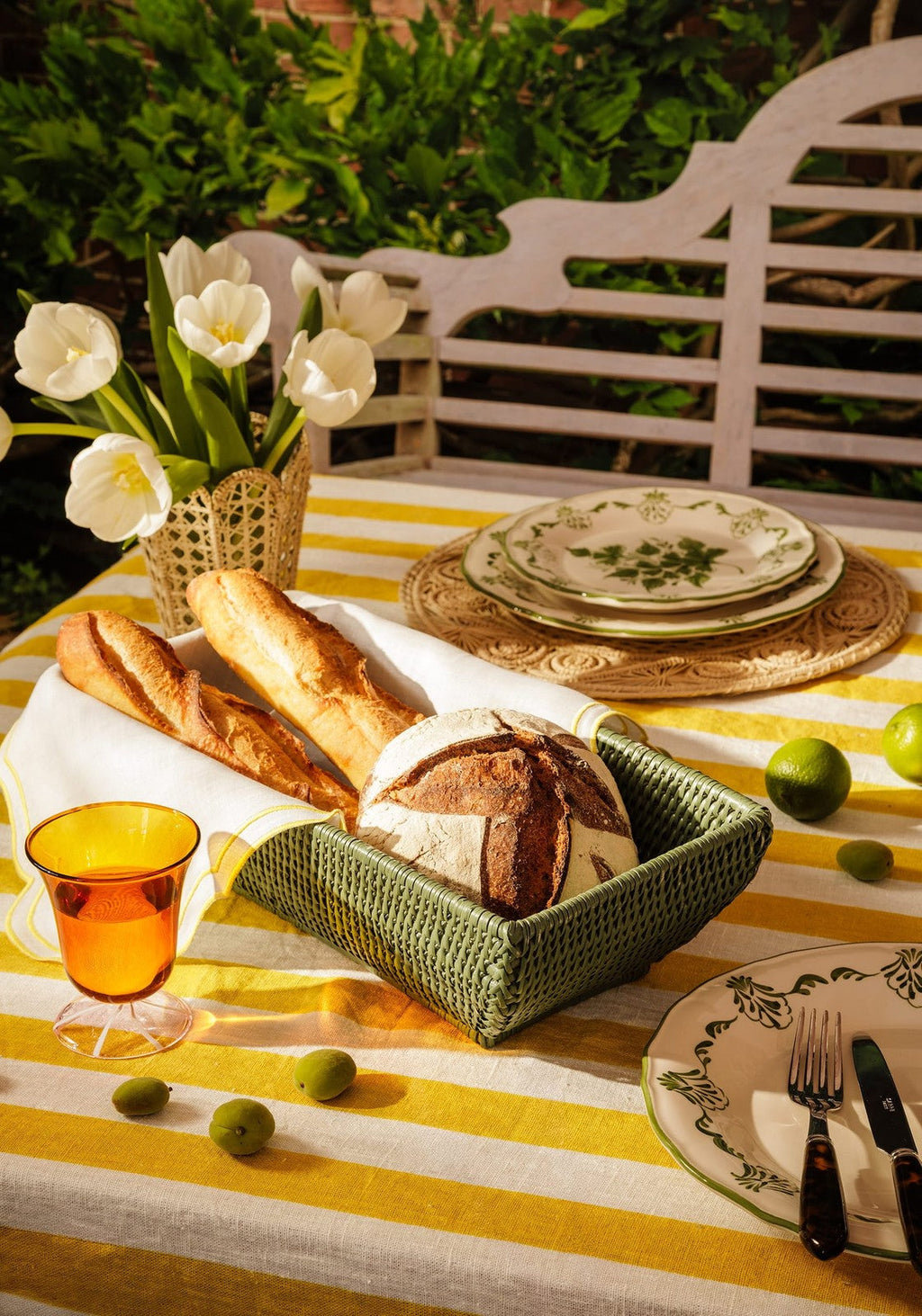 Outdoor table setting with bread, plates, and a vase of flowers on a yellow and white striped tablecloth.